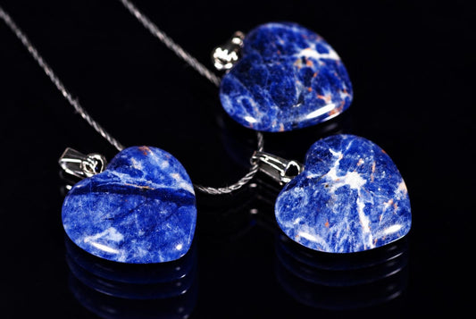 Three polished blue heart pendants with white markings and metal loops are arranged with black cords on a reflective black surface.