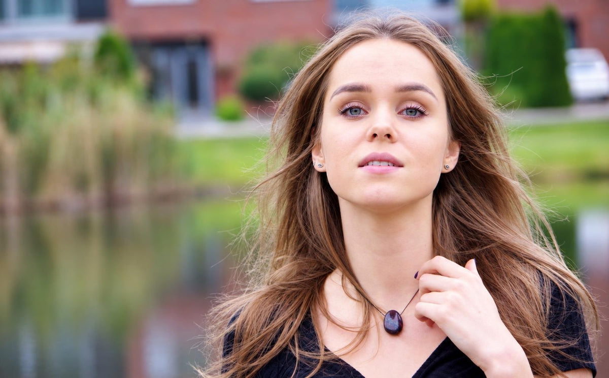 A woman wears a dark oval pendant on a thin cord around her neck, set against an outdoor background with water and greenery.