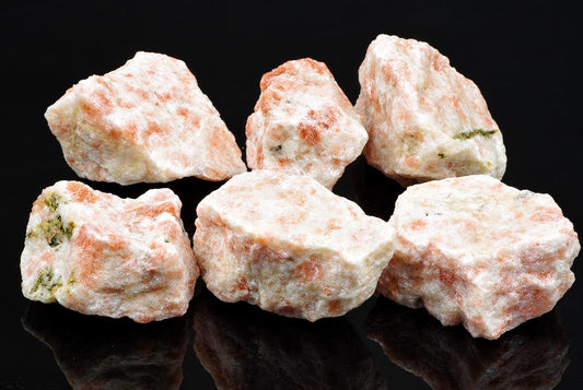 Six raw sunstone specimens with white and orange hues, some showing green inclusions, arranged on a dark reflective surface.