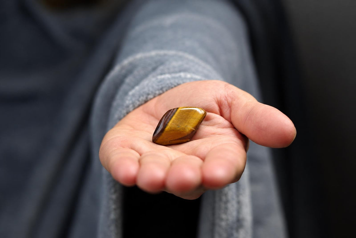 A hand holding a polished Tiger eye stone with golden and brown bands, showcasing the natural crystal’s smooth texture and reflective surface.
