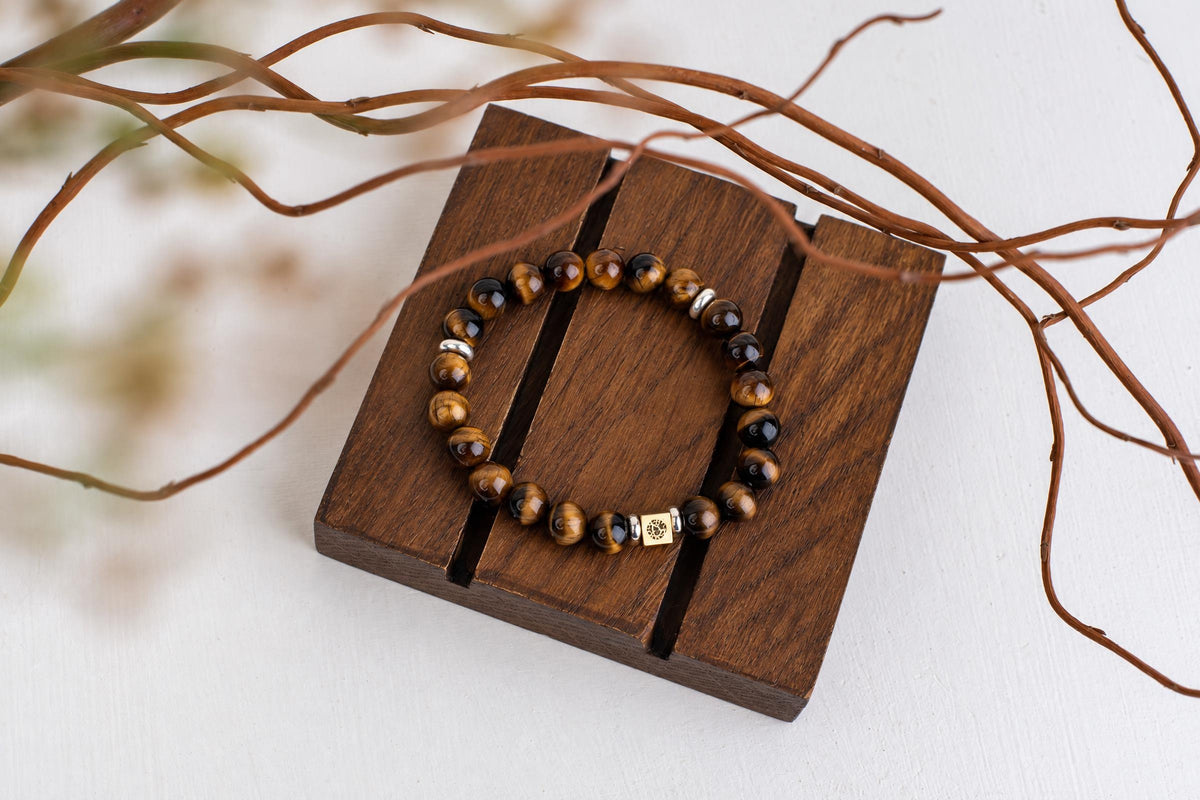 A beaded bracelet with tiger eye stones and a gold-toned square charm rests on a dark wooden surface.