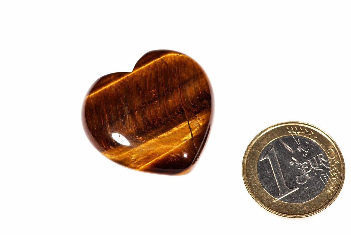 A polished heart-shaped tiger's eye stone with golden-brown stripes sits on a white surface next to a one-euro coin.