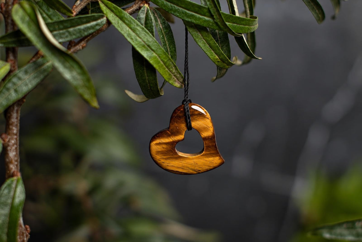 A polished, golden-brown heart-shaped pendant with a central cutout hangs from a black cord against green leaves.