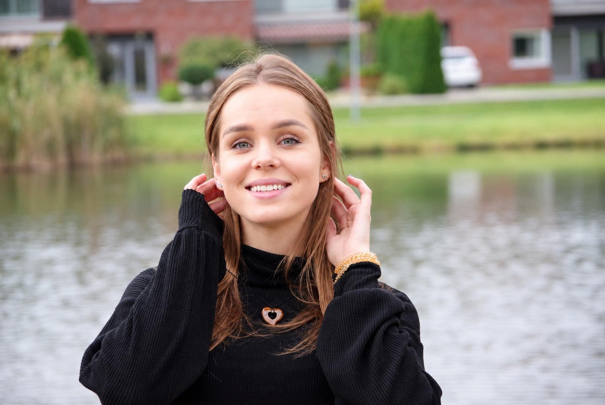 A woman wears a black turtleneck with a brown heart-shaped pendant necklace and gold-toned bracelet, smiling by water.
