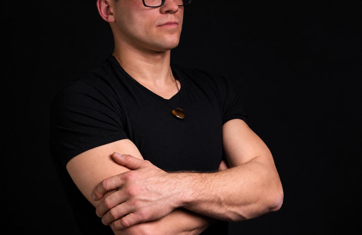 A man in a black shirt wears a dark brown, oval pendant on a thin cord against a dark background.