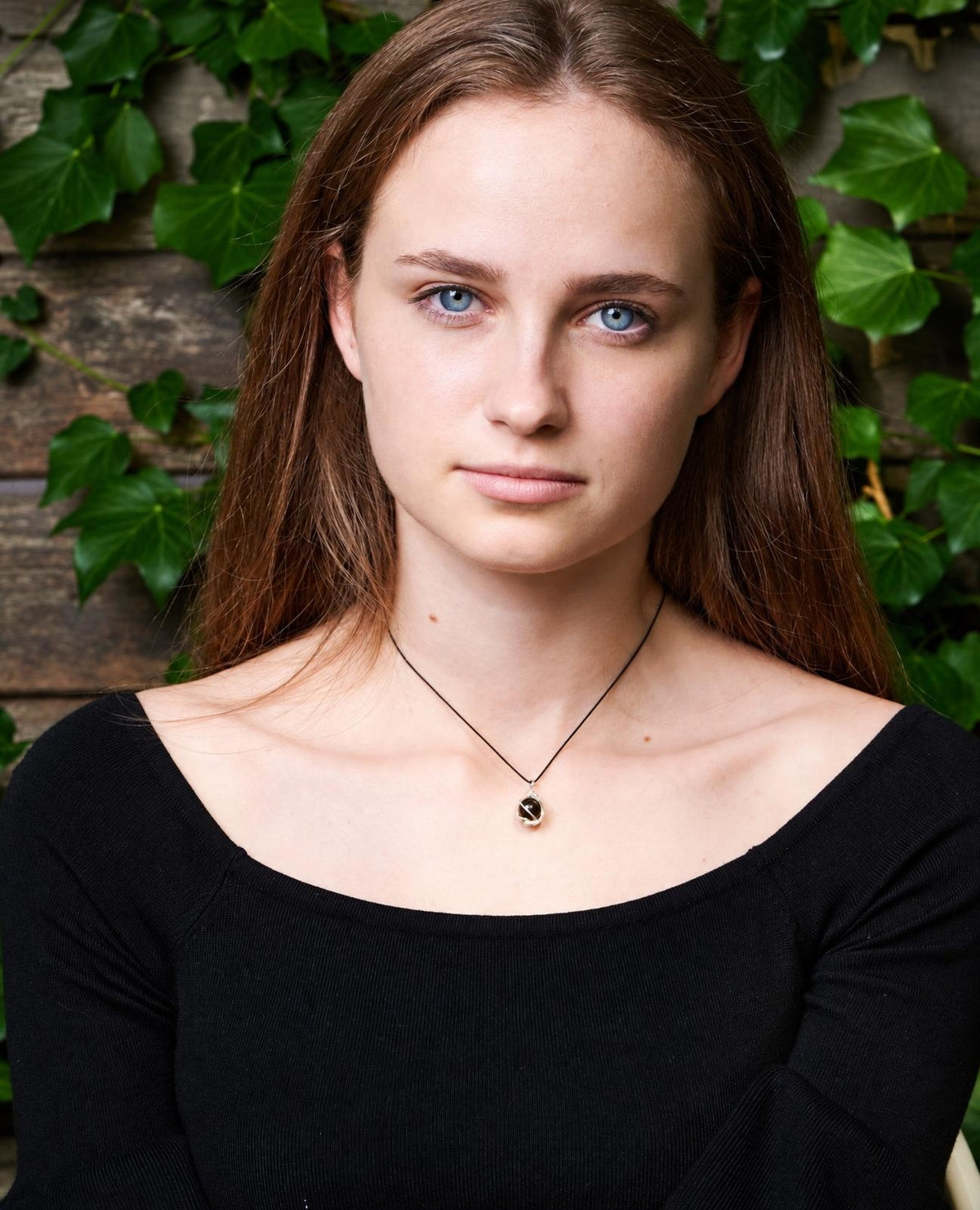 A woman wears a necklace with a small, dark sphere pendant on a thin black cord against a wooden background with green leaves.