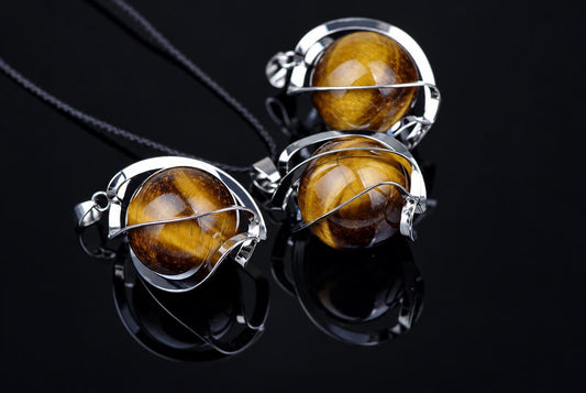 Three tiger eye sphere pendants with silver-toned wire settings on black cords, arranged on a dark reflective surface.