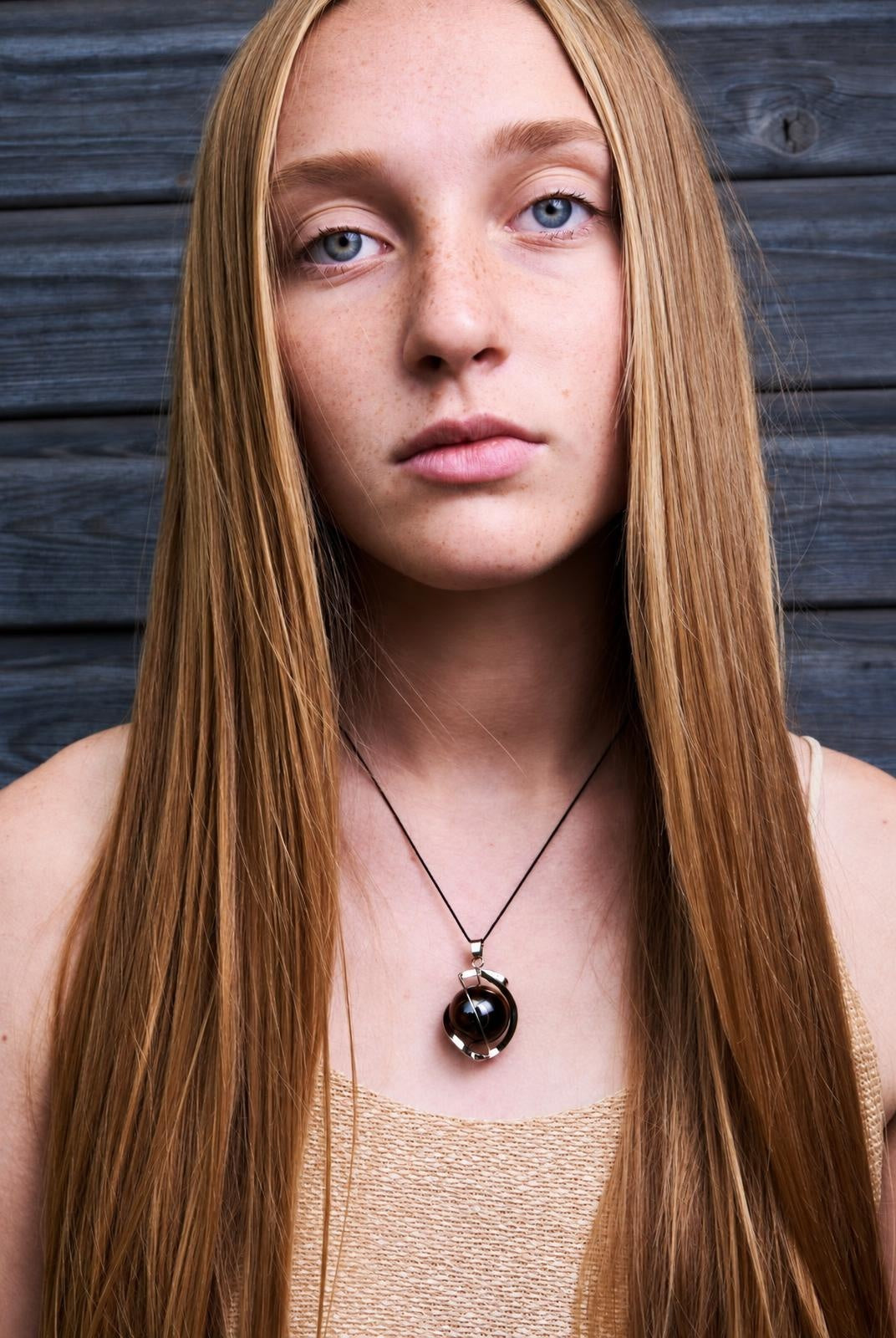 A woman wears a necklace with a dark brown sphere pendant set in a silver-toned ring on a black cord, against a dark wooden background.