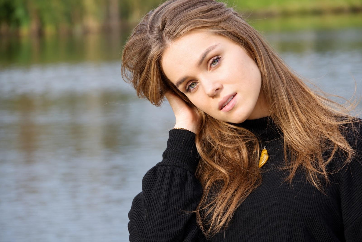 A woman with long brown hair wears a black top and a yellow pendant necklace against a blurred water background.