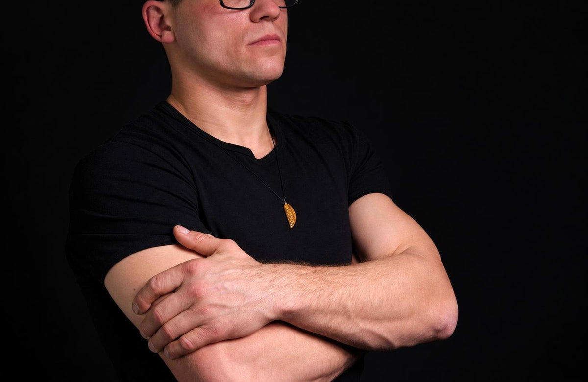 A man in a black t-shirt wears a gold-coloured wing-shaped pendant on a dark surface.