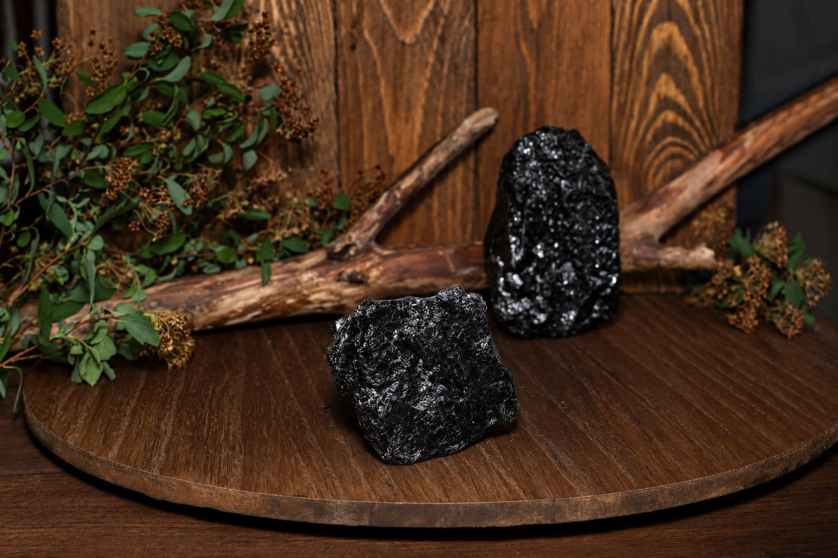 Two black, rough-textured stones sit on a dark wooden surface with greenery and branches in the background.