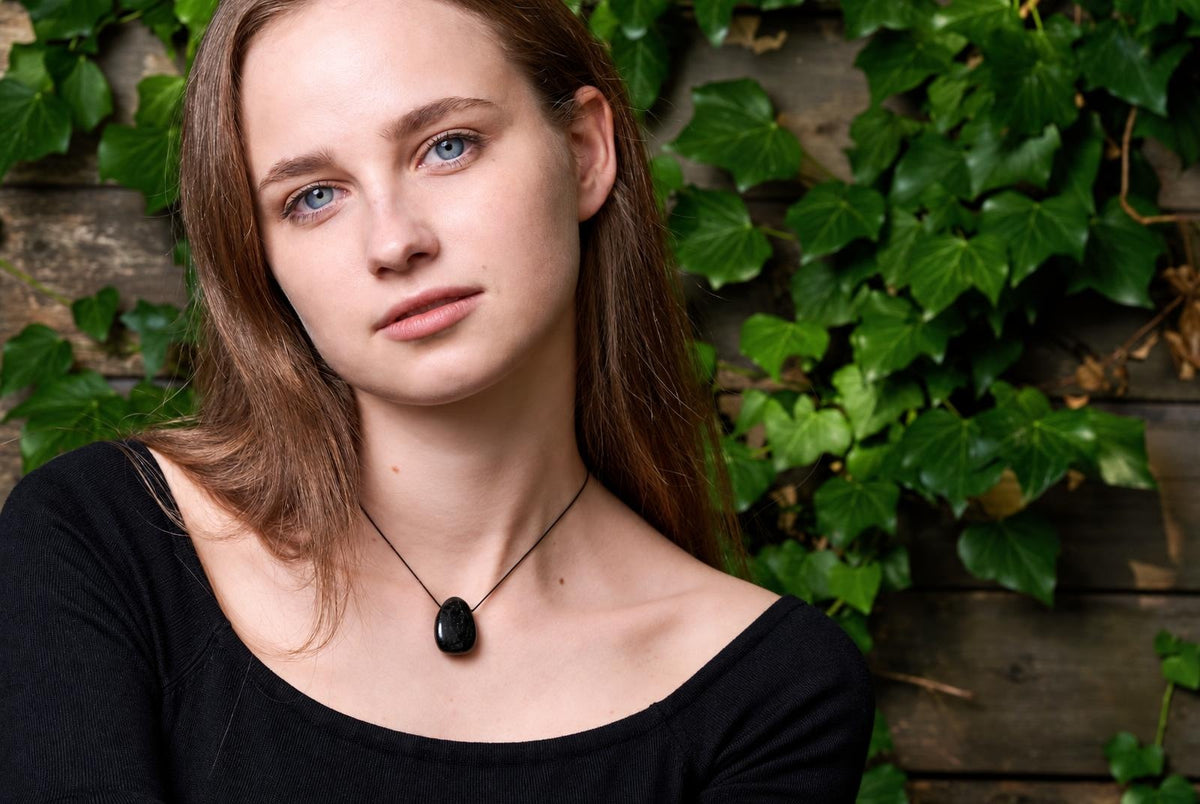 A woman wears a black pendant on a thin cord against a dark wooden surface with green ivy.
