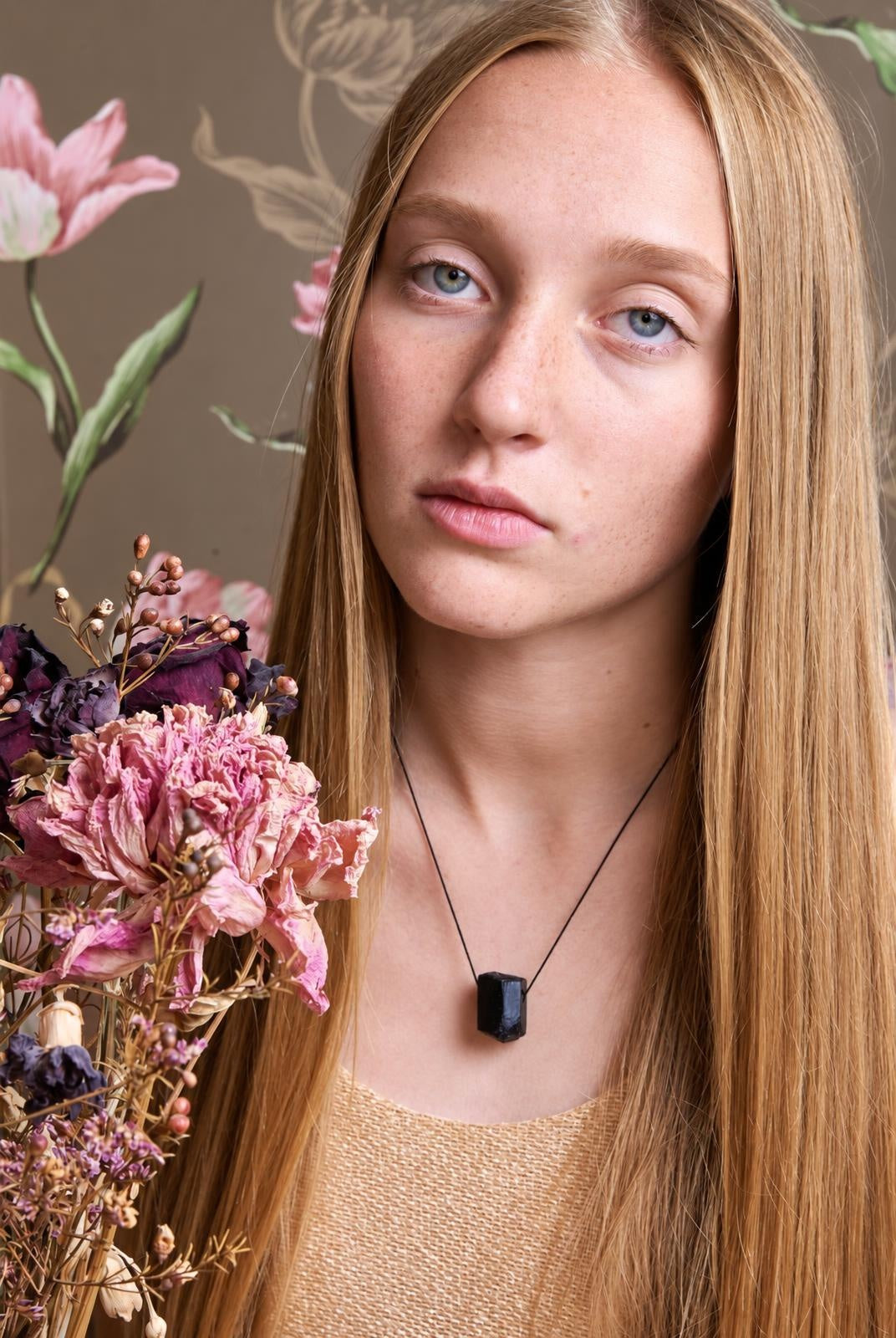 A woman with long blonde hair wears a black rectangular pendant on a thin cord, holding dried pink and purple flowers.