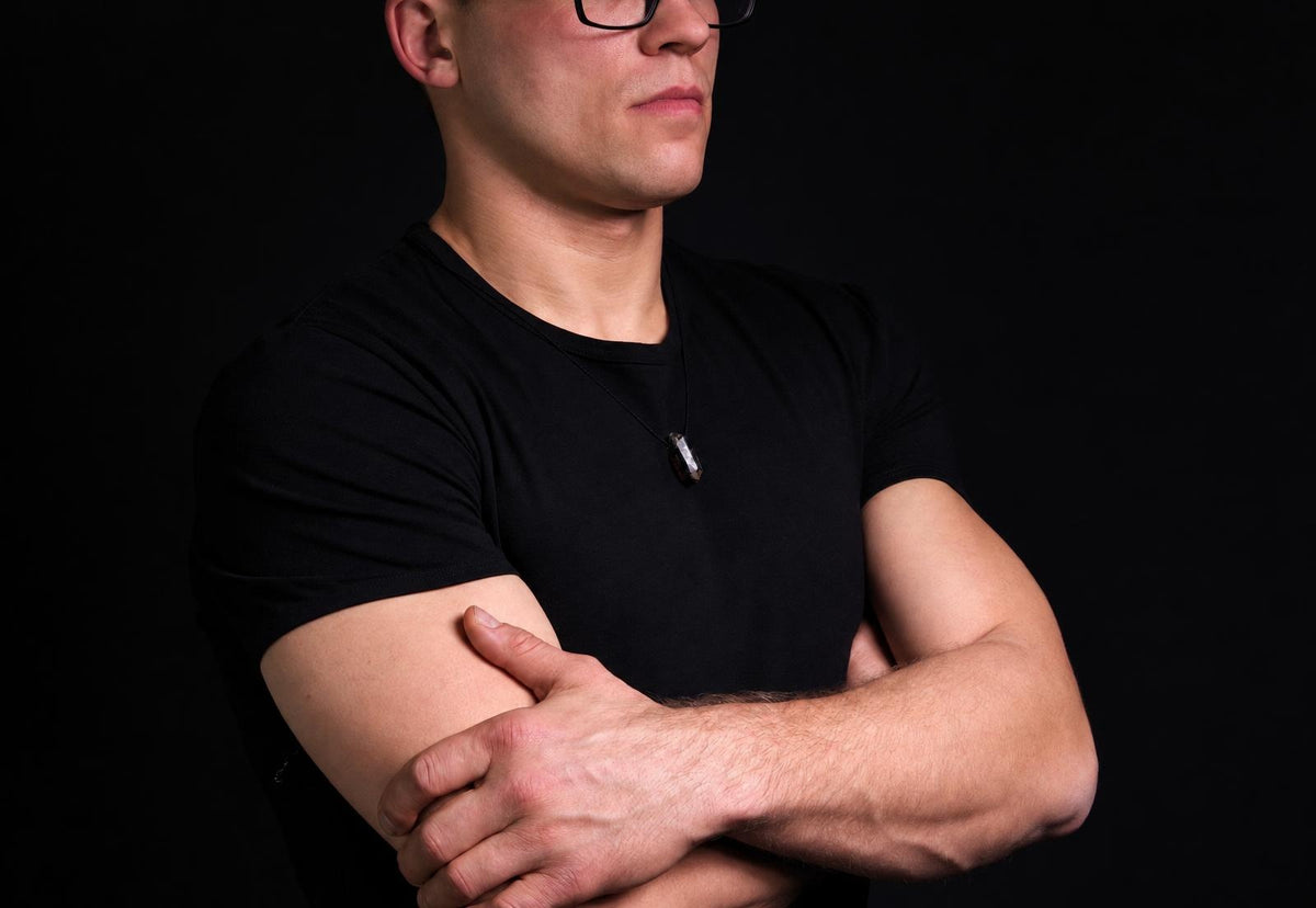 A man in a black t-shirt wears a dark, raw stone pendant on a thin cord against a dark background.