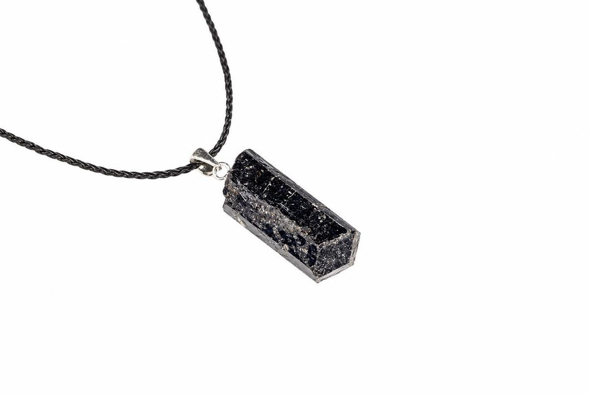 A dark elongated tourmaline pendant shows ridged texture within a silver-colored rectangular frame, suspended from a black cord on a white background.