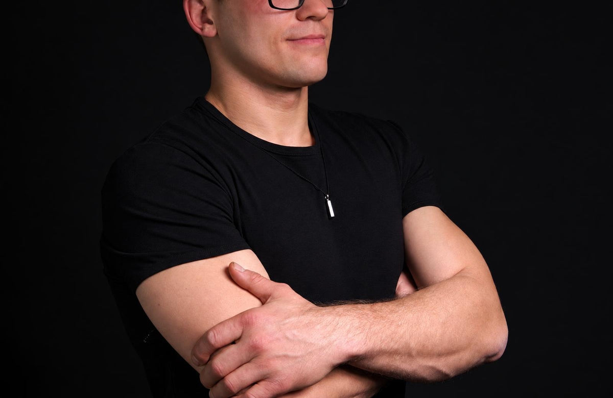 A man in a black t-shirt wears a silver-toned rectangular pendant on a thin cord against a dark background.