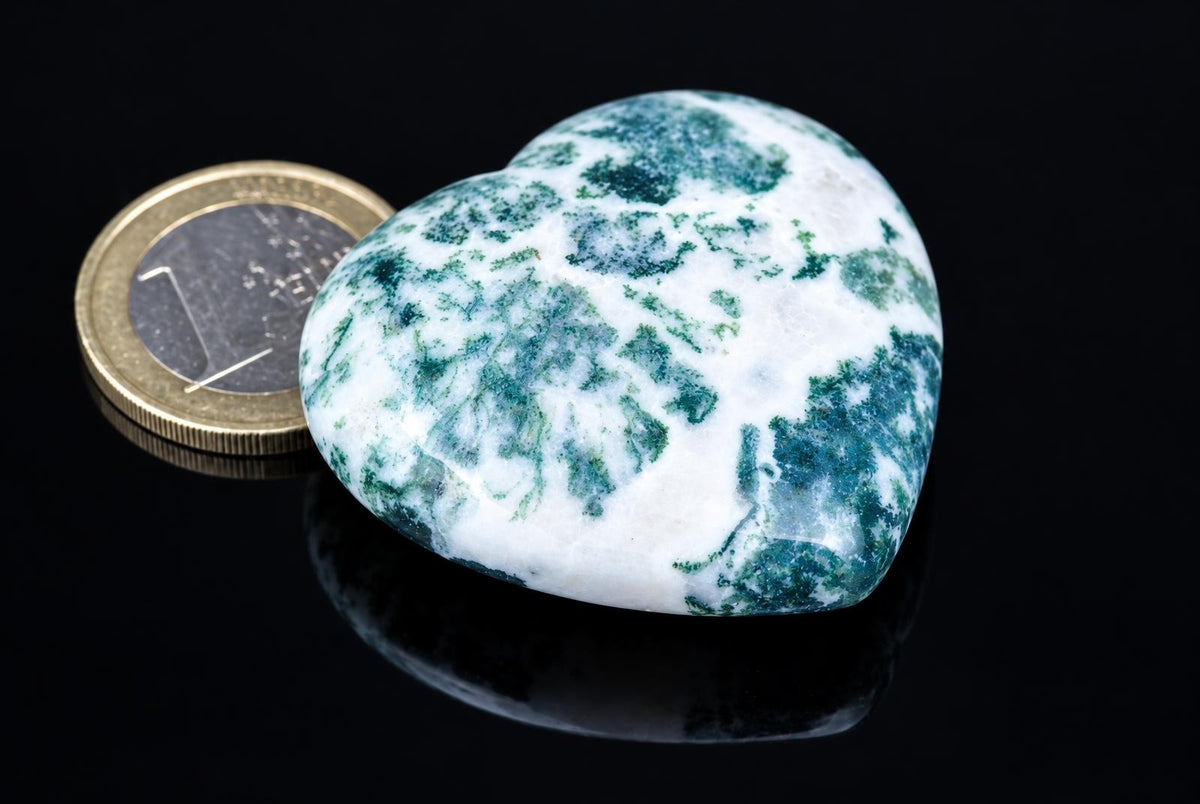 A polished heart-shaped stone with white and green patterns sits on a dark surface next to a coin.