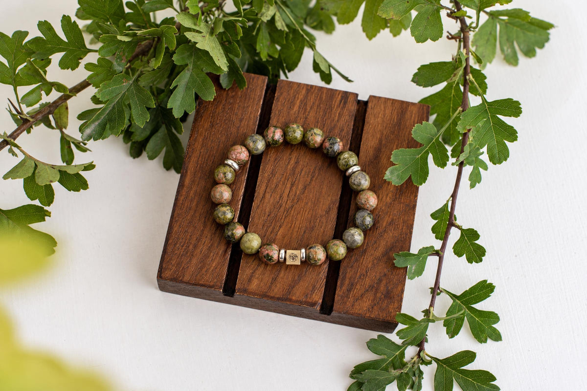 A beaded bracelet with green, pink and grey stones rests on a dark wooden stand, surrounded by green leaves.