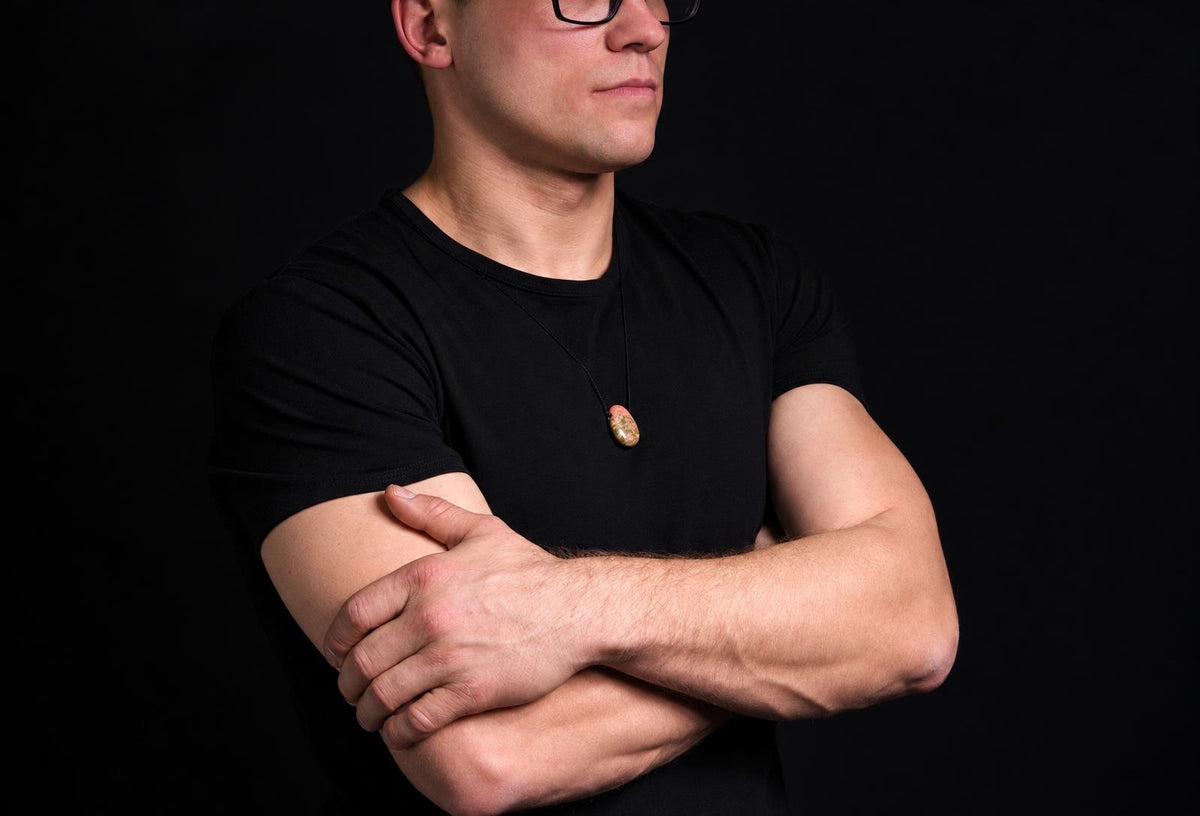 A man in a black t-shirt wears an oval pendant with green and pink tones on a dark surface.