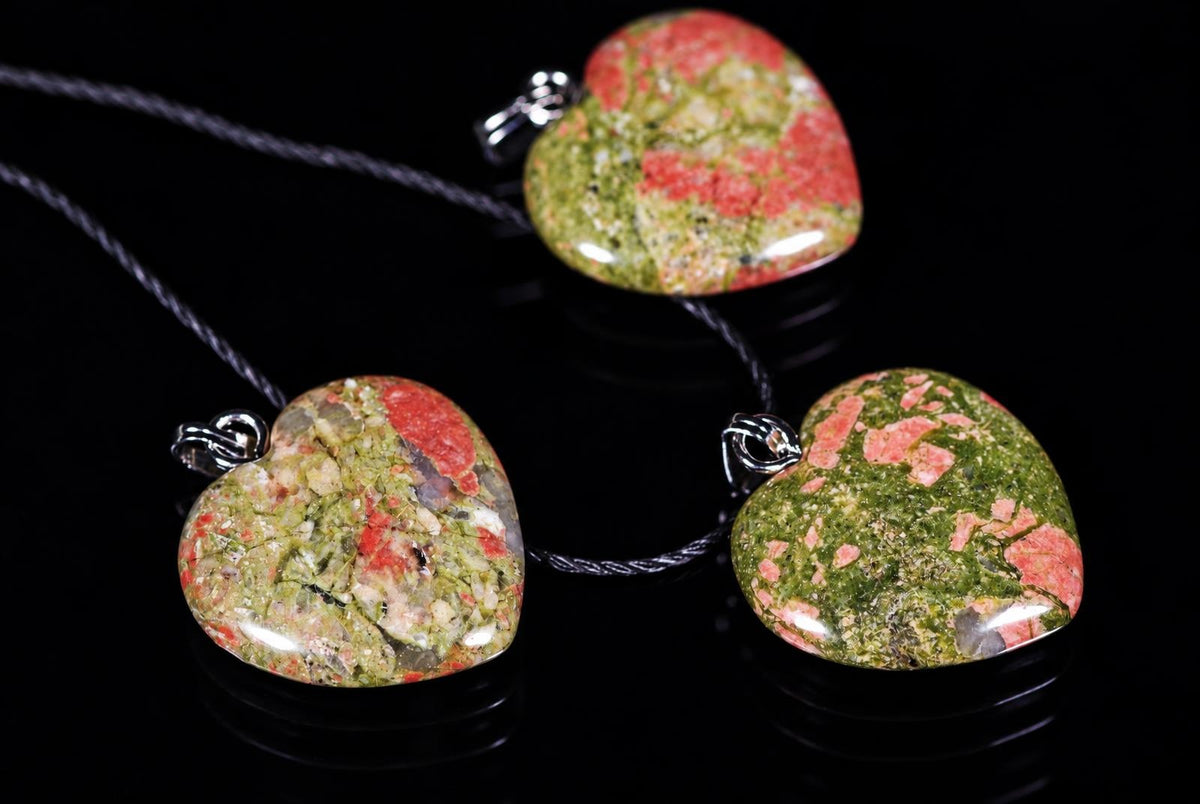 Three heart-shaped unakite pendants with green and red speckles on dark cords, resting on a black reflective surface.