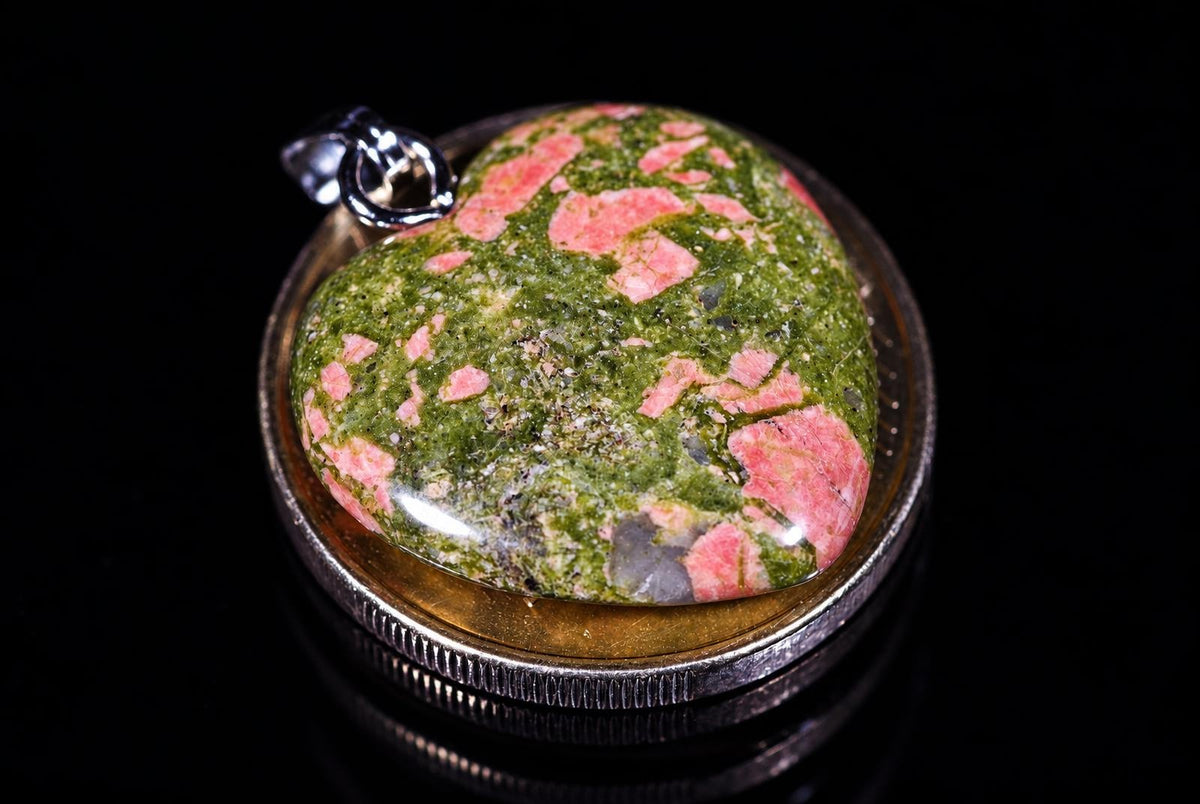 A heart-shaped unakite pendant with green and pink speckles set in a silver-toned bezel on a dark surface.