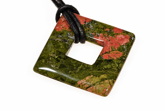 A square unakite pendant with a central hole, featuring green and red patterns, on a black cord against a white background.