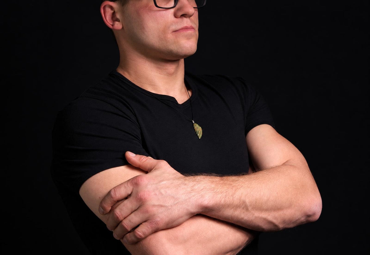 A man in a black t-shirt wears a pendant shaped like a wing, with green and brown tones, against a dark background.