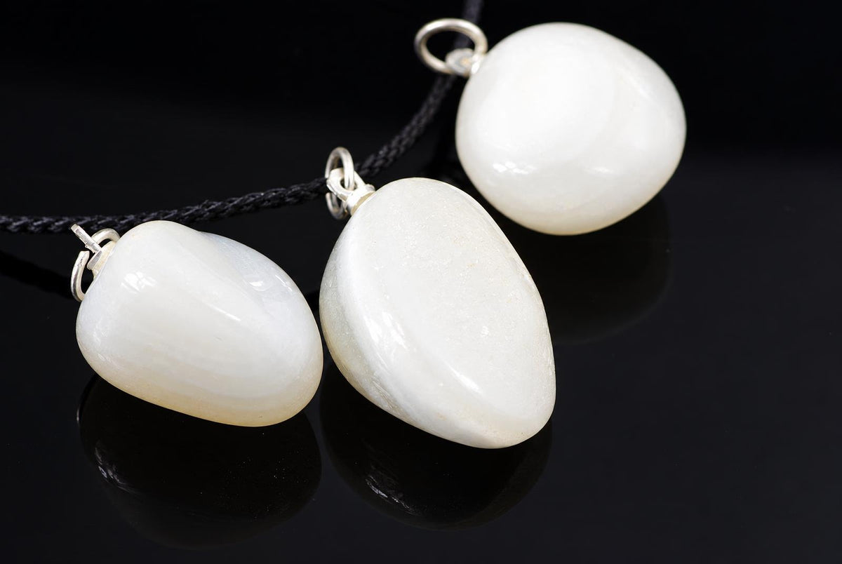 Three white pebble-shaped pendants with silver-toned loops on dark cords, resting on a dark reflective surface.