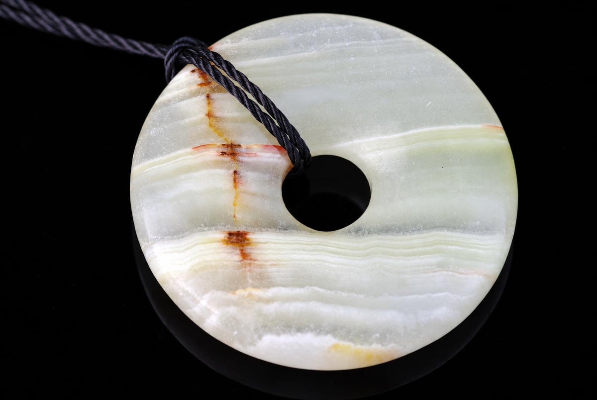 A round, donut-shaped pendant with a central hole, featuring white and pale green layers with brown streaks, on a dark surface.