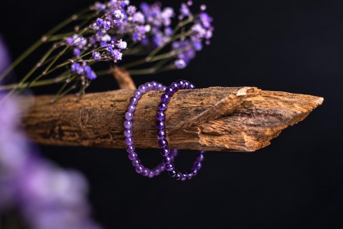 Two purple beaded bracelets hang over a weathered branch, with purple flowers against a dark background.