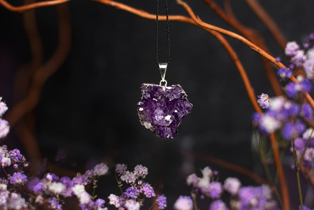 A purple crystal-cluster pendant hangs from a black cord among blurred purple flowers and brown branches.