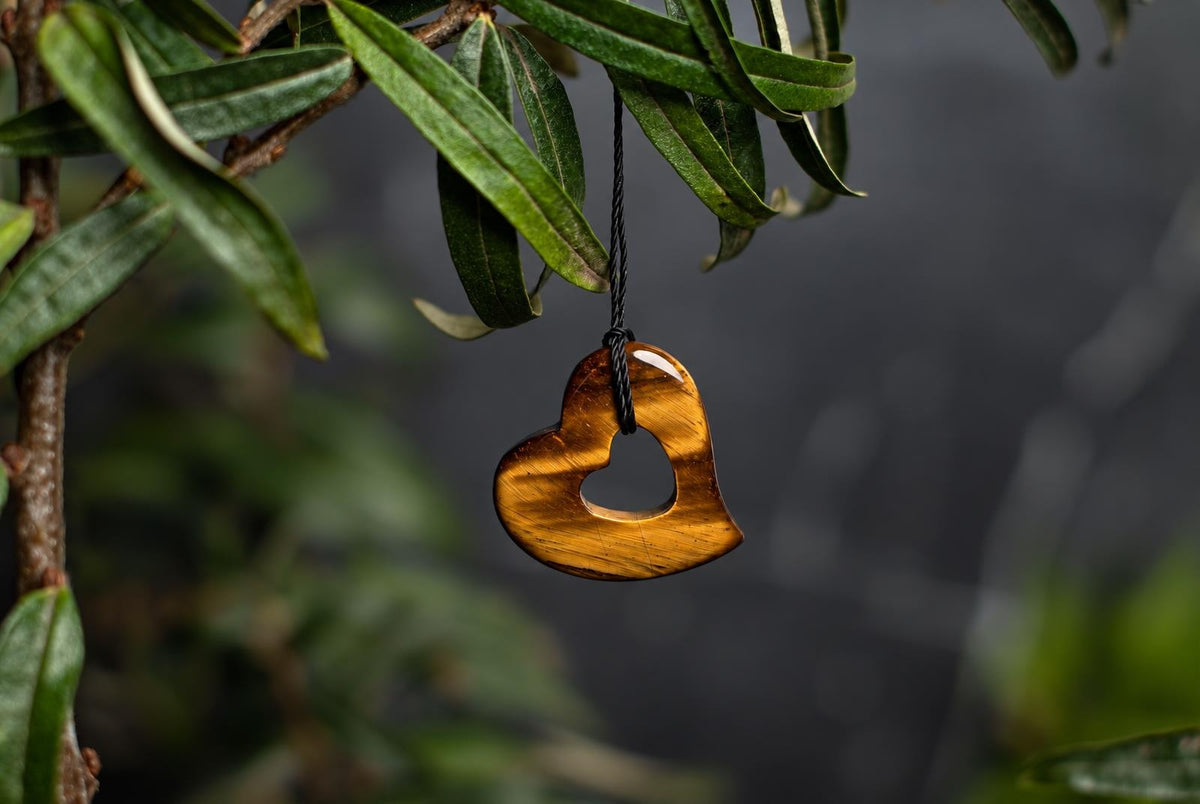 A polished golden-brown heart-shaped pendant with a smaller heart cutout, suspended by a black cord from green leaves.