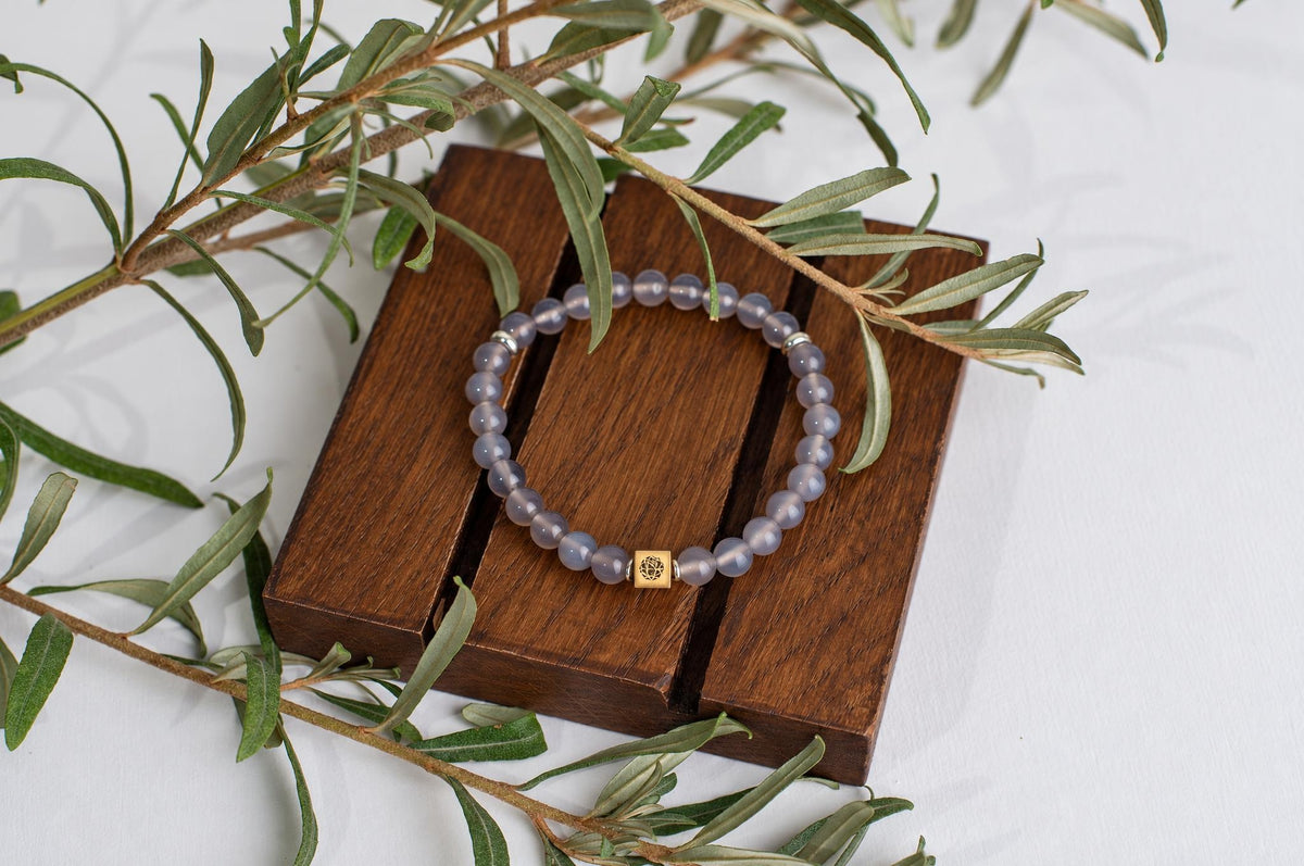 A bracelet with pale purple beads and a gold-toned square charm rests on a dark wooden surface, surrounded by green leaves.