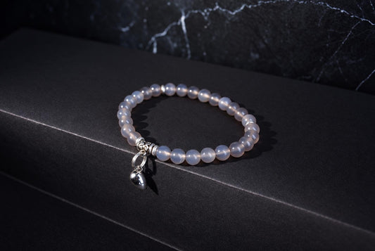 A bracelet with pale, round beads and a silver heart charm rests on a dark surface.