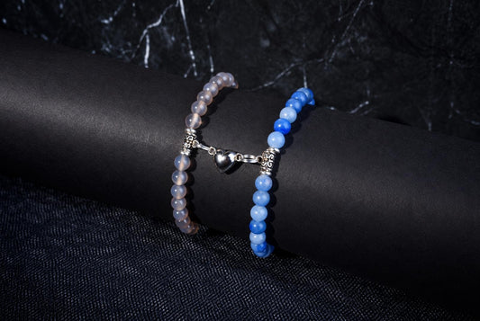 Two beaded bracelets on a dark surface: one with pale beads and a silver heart charm, the other with blue beads.
