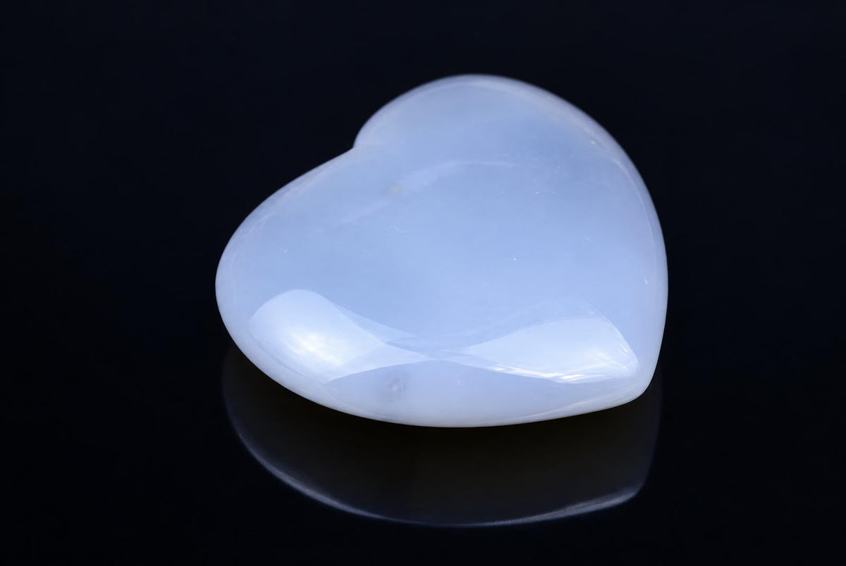 A polished, heart-shaped agate stone with a pale blue-white color rests on a dark surface.