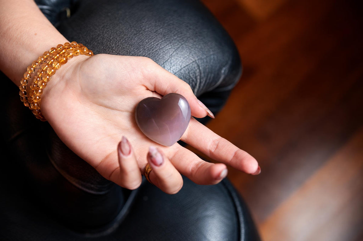 A hand holds a polished, heart-shaped stone with a smooth purple-to-pink gradient on a dark leather surface.