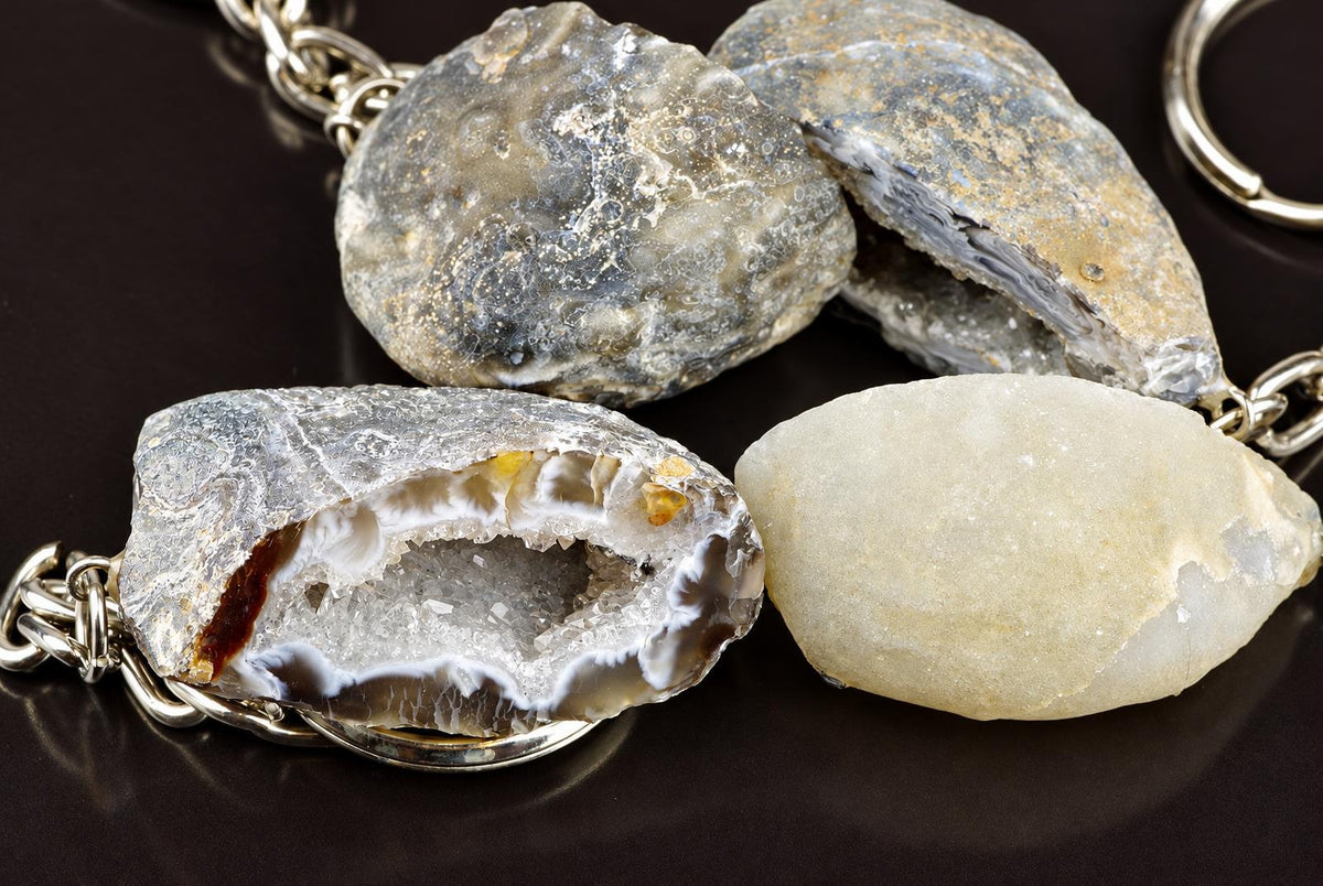 Four agate keychains with silver-toned chains on a dark surface. One is cut open, revealing white and brown crystals inside.
