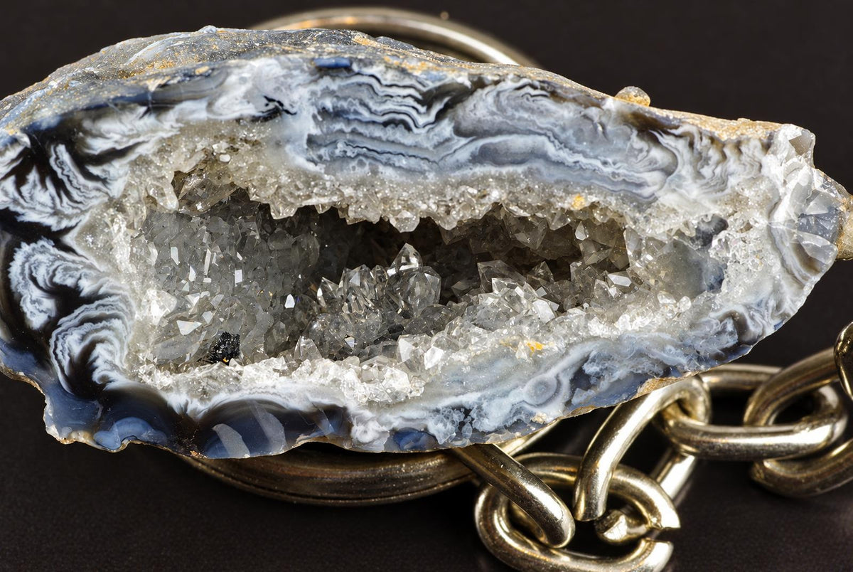 An agate keychain has grey and white bands surrounding a pale crystal cavity, with metal rings and a chain on a dark surface.