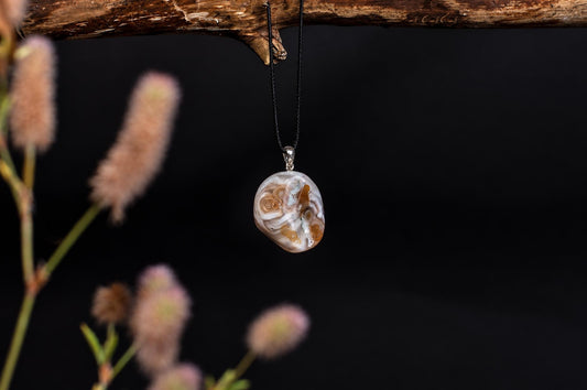 A polished agate pendant with white, brown and blue swirls hangs from a black cord on a dark surface.