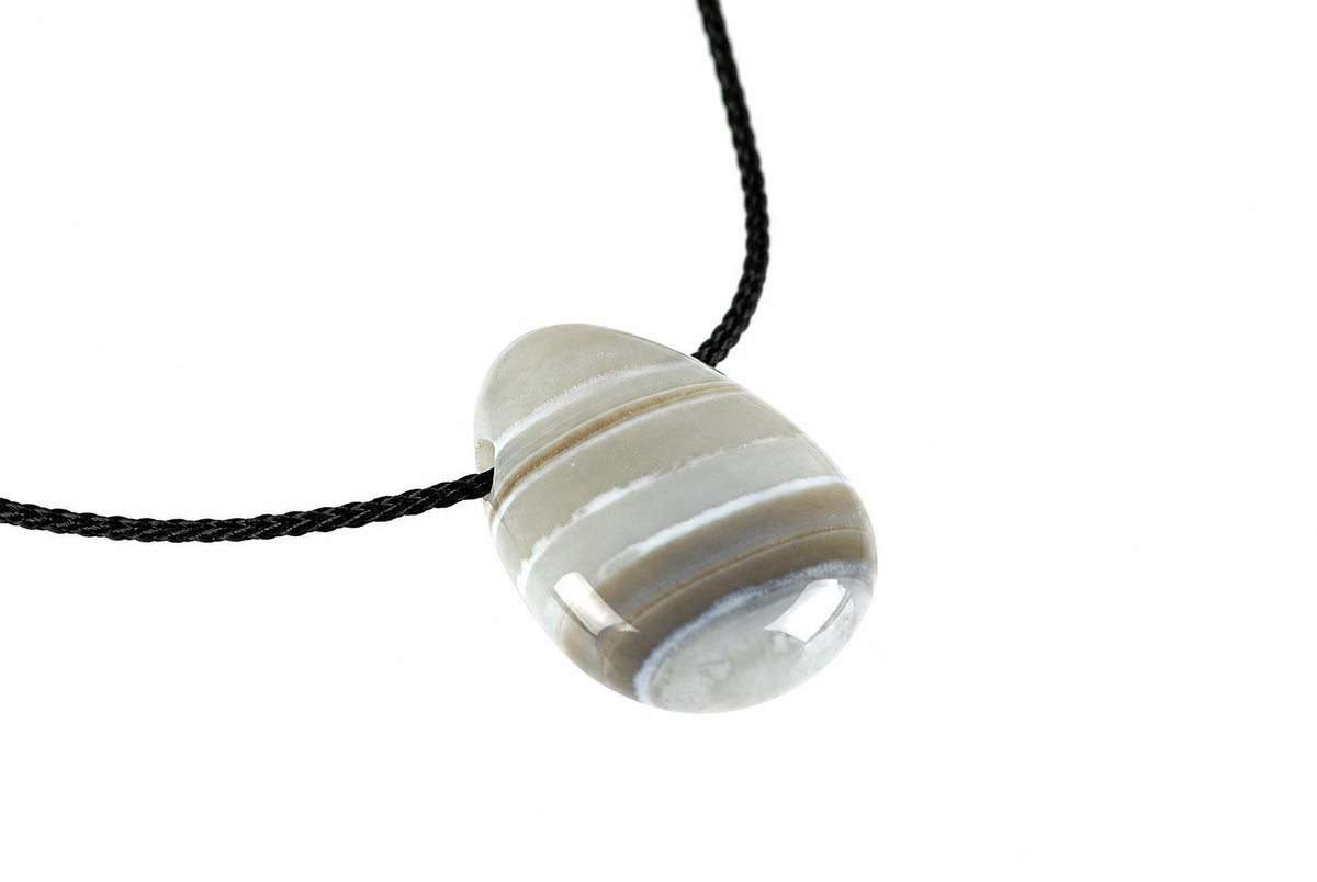 A polished agate pendant with horizontal bands of white, grey and brown hangs from a black cord against a plain white background.