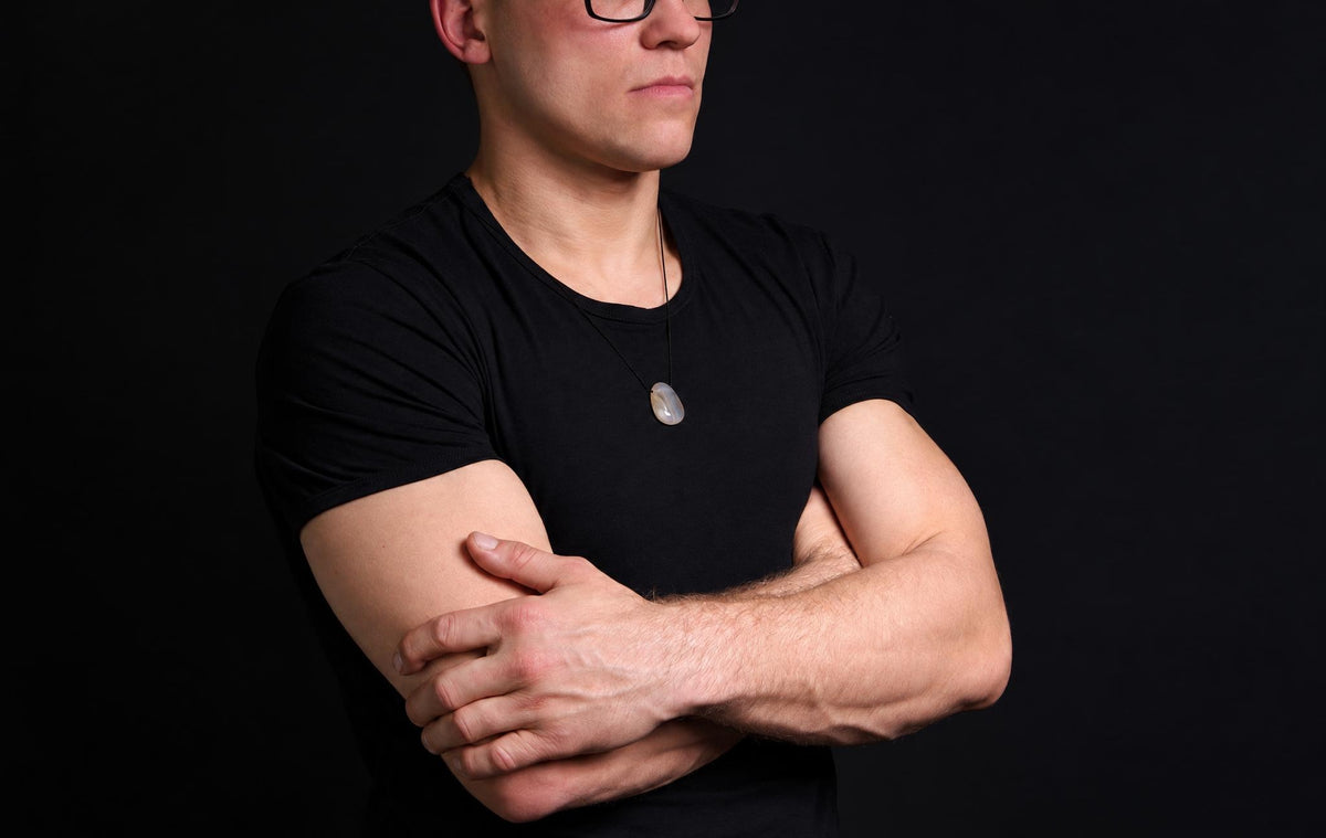 A man with folded arms wears a small pale agate drop pendant on a black cord over his black shirt.