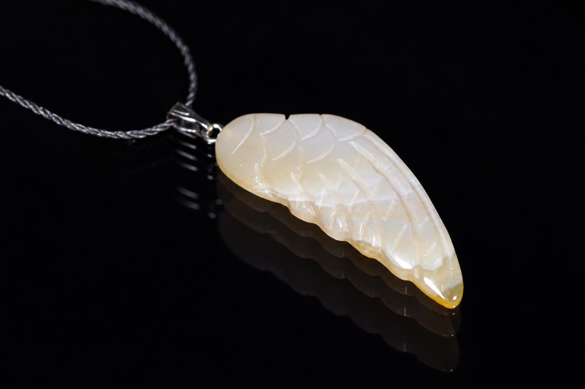 A silver-toned pendant with a carved, pale yellow and white wing-shaped stone hangs on a dark surface.