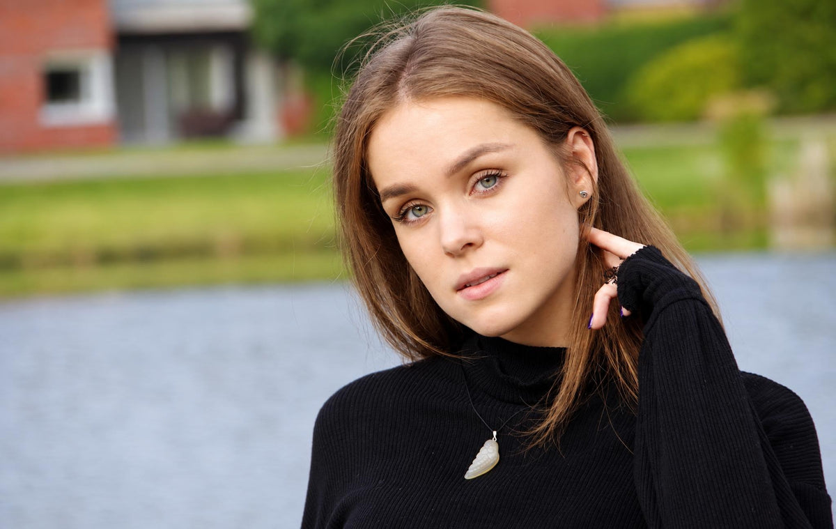 A woman wears a black turtleneck with a pale, wing-shaped pendant on a thin chain against a blurred outdoor background.