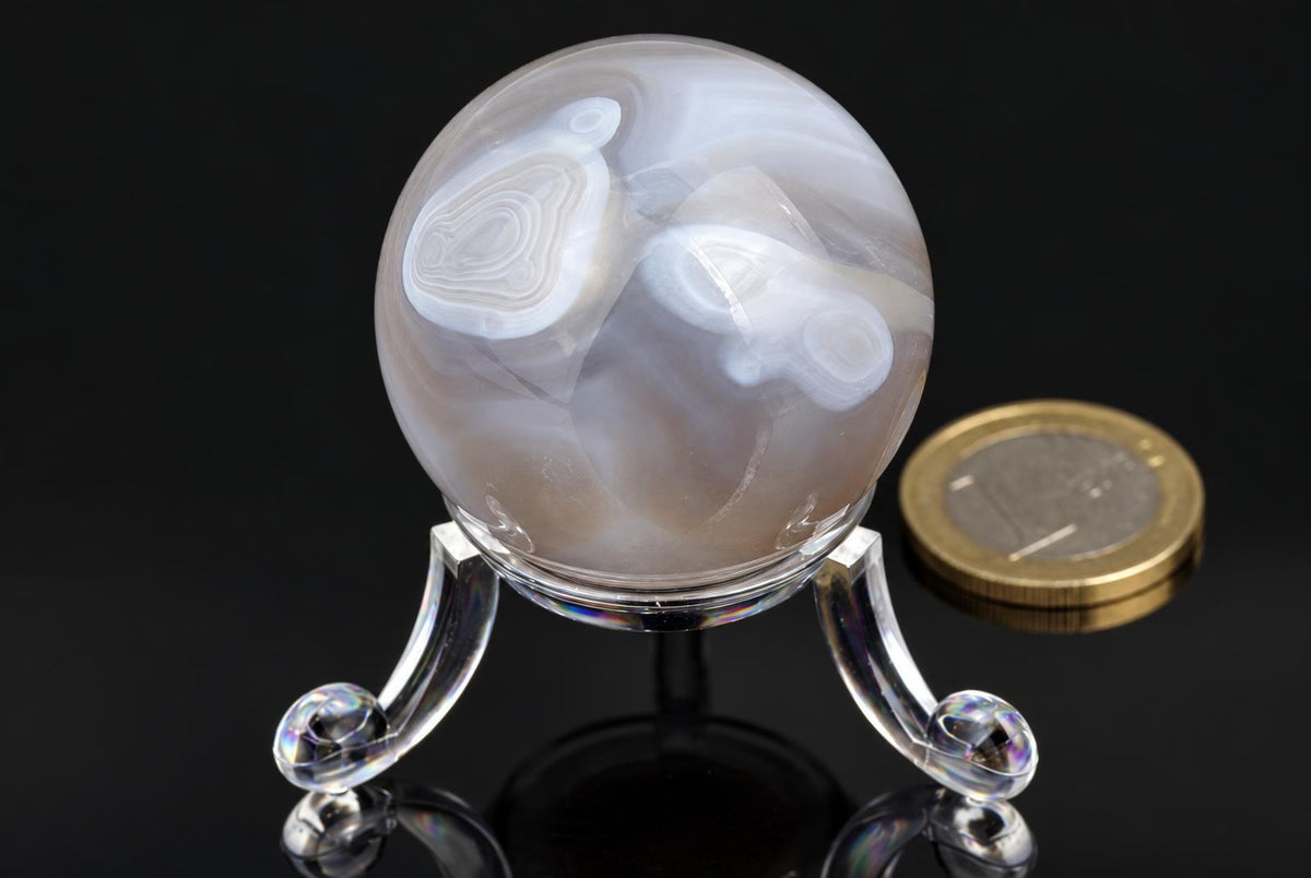 A polished agate sphere with white and brown swirls sits on a clear stand beside a coin on a dark surface.