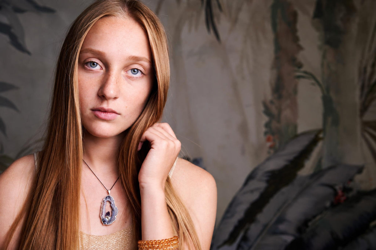 A woman wears a necklace with an irregular, dark grey and purple agate pendant on a thin cord.