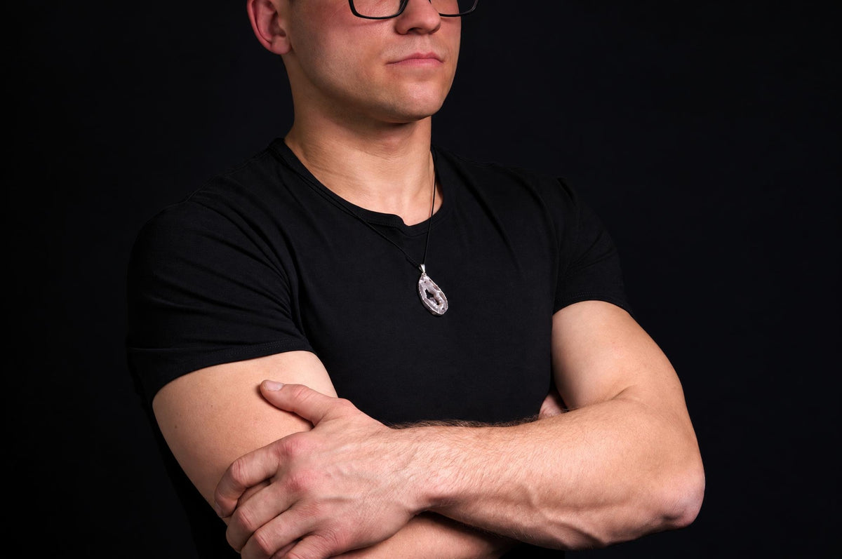 A man in a black t-shirt wears a silver-toned pendant with an irregular, translucent stone on a dark surface.