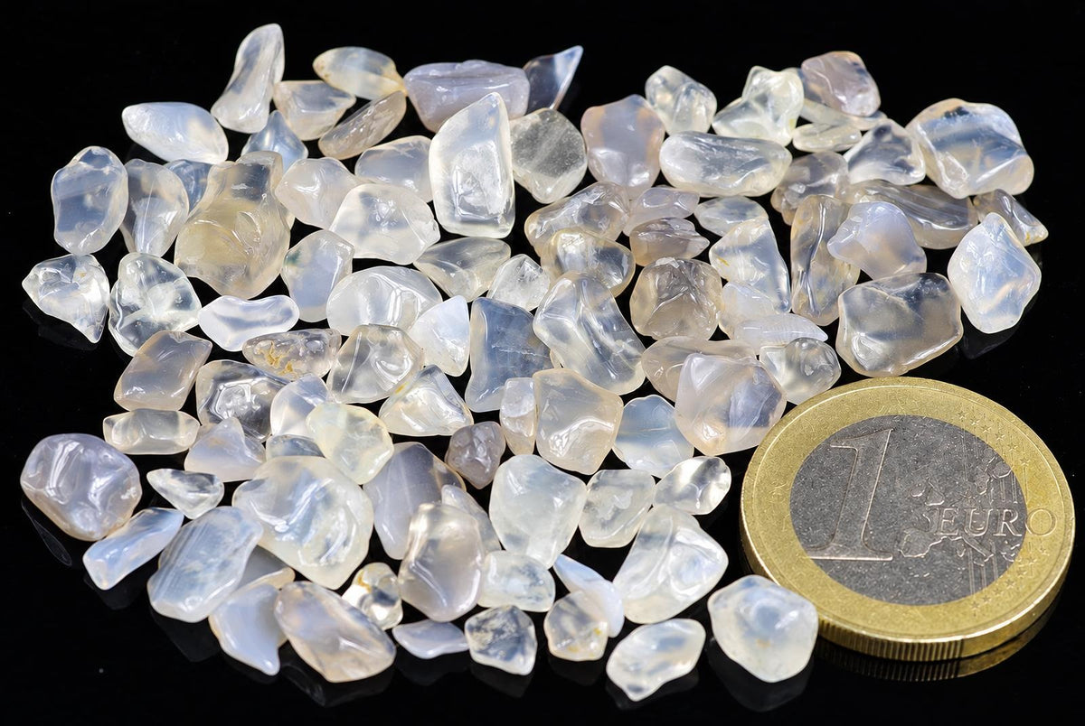A pile of small, translucent white and pale blue agate stones on a dark surface next to a one euro coin.
