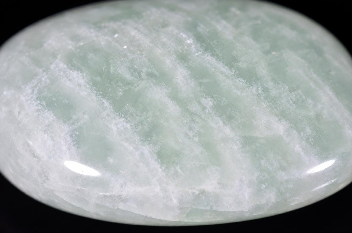 A polished, oval-shaped stone with a smooth surface. It features a mix of pale green and white swirling patterns against a dark background.