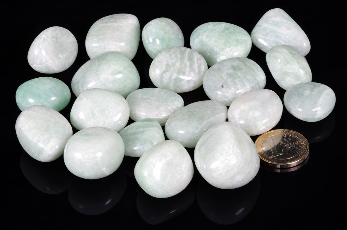 Polished amazonite stones show pale green and white colouring, gathered beside a coin on a glossy black surface with reflections.